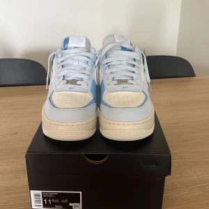 Nike Air Force 1 Shadow blå/vit helt nya  - Nike Air Force 1 Shadow i blått och vitt med snygga lager av ljusblått och krämvitt skinn. Klassisk AF1-sula, rund tå och coola detaljer som dubbla swooshes och broderad Nike-logga på hälen. Perfekt för dig som vill sticka ut med en unik sneaker.