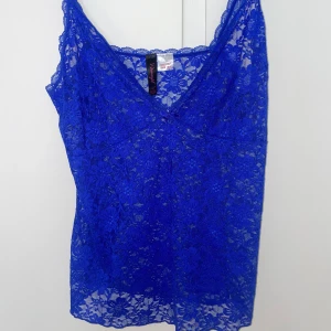 Blå spetslinne från dreamgirl  - Säljer ett blått linne från Dreamgirl i transparent spets med blommigt mönster. Linnet har smala axelband och v-ringning, perfekt för att ge en extra touch till din outfit. Superfint att styla lager på lager eller som det är.