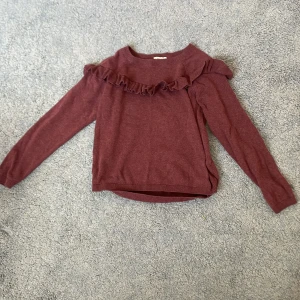 Vinröd tröja med volang från H&M - Vinröd långärmad tröja från H&M i storlek 134/140. Tröjan har en snygg volangdetalj över bröstet och axlarna, rund halsringning och ribbade muddar. Perfekt för dig som gillar en enkel men ändå söt stil.