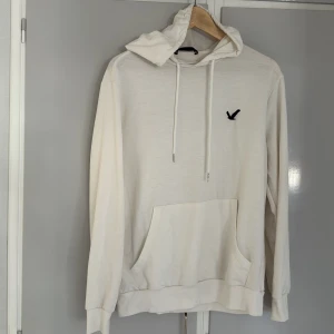 Vit hoodie från SHEIN med broderad logga - Vit hoodie från SHEIN med svart broderad logga på bröstet. Tröjan har huva med snörning, stor känguruficka framtill och långa ärmar. Perfekt för en chill och avslappnad stil. Materialet känns mjukt och skönt mot huden.