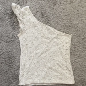 Vit spetsig oneshoulder topp - Superfin vit topp med spets och virkat mönster framtill. Toppen är oneshoulder med en axel täckt av spetsdetaljer och resten av toppen är slät baktill. Perfekt för dig som gillar unika detaljer och somrig stil.