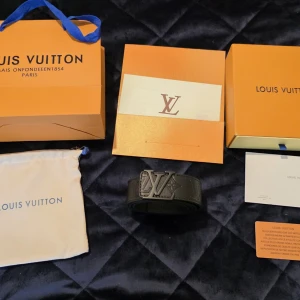 Svart Louis Vuitton skärp med LV-spänne - Snyggt svart skärp från Louis Vuitton med ikoniskt LV-spänne i svart metall. Bältet har en stilren design med diskret mönster i lädret och bred rem. Perfekt accessoar för att lyfta din outfit med lyxig känsla.