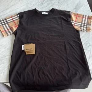 Svart Burberry t-shirt med rutiga ärmar - Svart t-shirt från Burberry med klassiska beige och rödrutiga ärmar. Ikoniskt mönster på ärmarna och enkel rund halsringning. Skön bomullskvalitet och snygg passform som sticker ut.