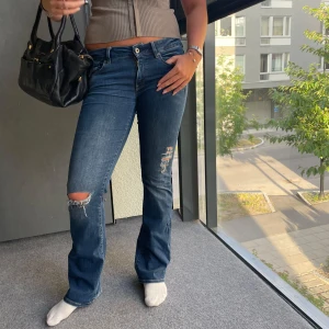 Blå bootcut jeans med slitningar - Snygga blå jeans  bootcut passform och slitna detaljer på knä och lår. Jeansen har låg midja och klassisk femficksdesign. Perfekta för dig som gillar en avslappnad och trendig look.