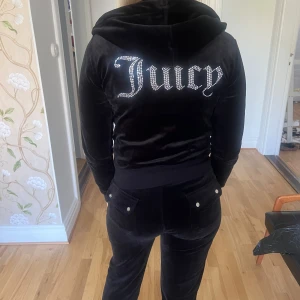 Juicy Couture svart velourjacka XS - Svart velourjacka från Juicy Couture med huva, dragkedja och glittrande 'Juicy'-text på ryggen. Jackan har långa ärmar, ribbade muddar och en snygg croppad passform. Perfekt för dig som vill ha den ikoniska 2000-talslooken.