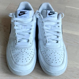 Nike Court Vision vita sneakers - Fräscha Nike Court Vision sneakers i vitt skinn med klassisk svart swoosh på sidorna. Skorna har perforerad tå, vit platt sula och snörning. Perfekt för dig som gillar en clean och sportig look med retrovibbar.