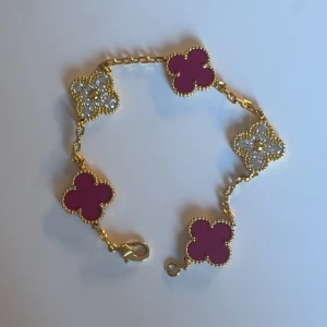 Guldarmband med rosa och glittriga blommor - Superfint armband i guldton med fyra rosa fyrklöver och två glittriga blommor med stenar. Kedjan är tunn och detaljerad, och varje blomma har en pärlad kant. Perfekt accessoar för att lyfta din stil med färg och bling.