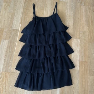 Svart volangklänning från Lindex Kids - Supersöt svart kortklänning från Lindex Kids med flera lager volanger och smala axelband. Klänningen har dragkedja i ryggen och är tillverkad i ett lätt, flowy material som ger en luftig känsla. Perfekt för dig som gillar en lekfull och trendig stil. Finns inte längre kvar i sortimentet på Lindex. Köparen betalar för frakt.