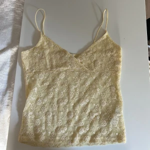 Gul spetslinne från Subdued - Supersött ljusgult linne från Subdued med tunna axelband och v-ringning. Hela toppen är täckt av blommig spets och har en mjuk, feminin vibe. Perfekt att styla med jeans eller kjol för en trendig look. 