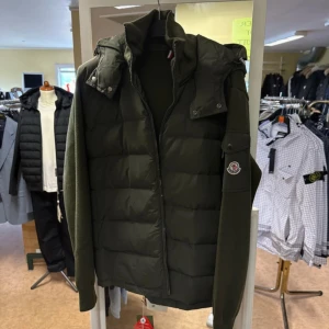 Grön dunväst från Moncler - Snygg grön dunväst från Moncler med quiltad front och stickade ärmar. Västen har dragkedja, hög krage och avtagbar huva. Klassisk Moncler-logga på ärmen och diskret ribbad detalj. Perfekt för lager-på-lager och streetstyle.
