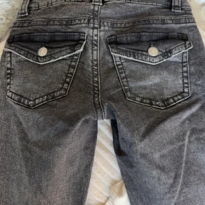Gråa bootcut jeans med fickdetaljer - Säljer ett par gråa bootcut jeans med snygga ficklock och silverfärgade knappar bak. Jeansen har låg midja och klassisk femficksdesign. Perfekta för dig som gillar en avslappnad men trendig stil.