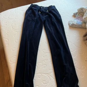 Mörkblå velourbyxor Juicy Couture XS - Mörkblå mjukisbyxor från Juicy Couture i velour med snörning i midjan och fickor både fram och bak. Klassisk bootcut-modell med snygga detaljer och guldiga metalldelar på snörena. Perfekta för en chill och trendig look.