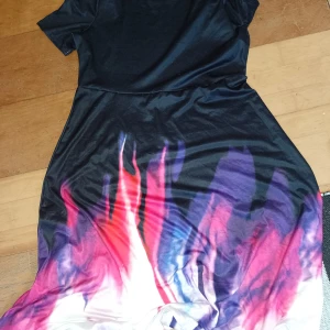 Svart klänning med färgstark print - Cool svart klänning med korta ärmar och färgstarkt mönster i rött, lila och vitt nedtill. Mjuk och glansig känsla, perfekt för dig som gillar att sticka ut. Sitter snyggt och har en avslappnad passform.