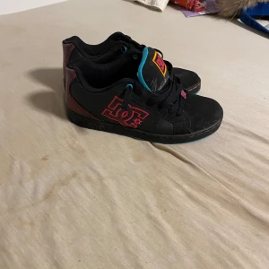 Svarta DC sneakers med rosa logga - Svarta sneakers från DC med stor rosa logga på sidan och detaljer i blått och gult. Skorna har snörning och platt sula, samt inslag av rött på hälen. Perfekta för dig som gillar streetstyle och skate.