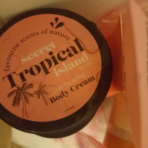 Tropical Island Body Cream - En mjukgörande body cream med doft av tropisk ö. Kommer i en rund burk med svart lock och persikofärgad etikett med palmer. Perfekt för dig som vill ha en fräsch och exotisk känsla på huden.