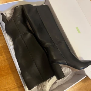 Svarta höga stövlar med klack - Köpta från NA-KD, äkta läder.   Dem är köpta för 1499kr, men säljer för 1000kr. Dem är inte använda.                               Stilrena svarta stövlar i skinn med spetsig tå och bred klack. Snygg siluett med dragkedja på insidan för enkel påtagning. Perfekta till både kjol och jeans för en trendig look.