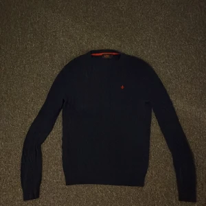 Mörkblå stickad tröja från Morris  - Mörkblå stickad tröja från Morris med klassisk rund hals och diskret röd logga broderad på bröstet. Tröjan har lång ärm och ribbade muddar. Perfekt för dig som gillar stilren och tidlös design. Storlek S