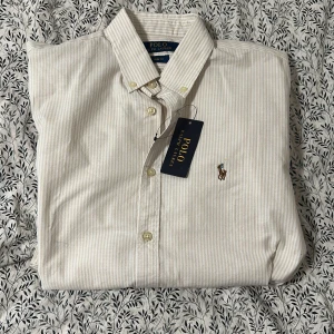Randig skjorta Polo Ralph Lauren M - Klassisk slim fit skjorta från Polo Ralph Lauren i beige och vit med tunna ränder. Skjortan har button down-krage, broderad logga på bröstet och långa ärmar. Tillverkad i mjuk bomull och stängs med knappar framtill. Ej använd 