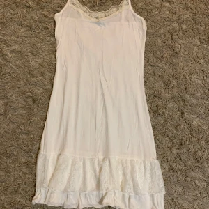 Vit spetsklänning med volangkant - Söt vit kortklänning med smala axelband och spetsdetaljer vid halsringningen. Klänningen har en volangkant nertill med blommig spets och är perfekt för sommarens varma dagar. Mjuk och lätt i materialet, ger en romantisk och avslappnad vibe.