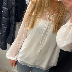 Vit prickig blus med knytband - Säljer en söt vit blus med transparenta, långärmade ärmar och diskret prickigt mönster. Blusen har knytband vid halsen och fina spetsdetaljer vid ärmsluten. Luftig och flowy passform som ger en romantisk vibe.