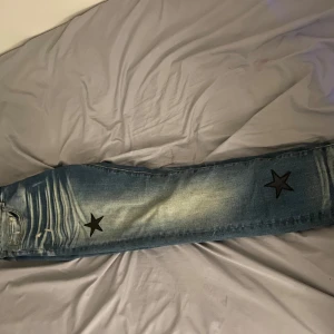 Amiri byxor  - Snygga blå jeans med slitningar och coola svarta stjärnpatches på benen. Jeansen har en klassisk femficksmodell, normal passform och är tillverkade i denim. Perfekta för dig som vill sticka ut med en unik detalj på dina byxor. Köpta från företag 