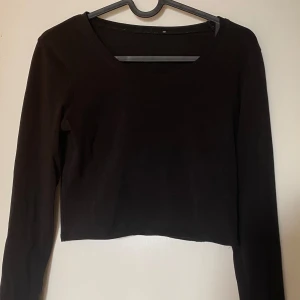 Svart croppad långärmad topp - En svart, enkel och croppad långärmad topp med rund halsringning. Perfekt att matcha med höga jeans eller kjol för en clean look. Mjuk och stretchig känsla, passar dig som gillar minimalistisk stil. Frakt tillkommer