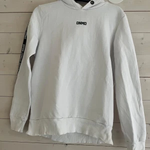 Vit hoodie från Denim Day med tryck - Vit hoodie från Denim Day med svart DNMD-tryck på bröstet och text längs ena ärmen. Tröjan har huva med metallöljetter och ribbade muddar. Perfekt för en chill streetwear-look. Materialet är mjuk bomull och modellen är normal i passformen.  Obs den är lite trasig vid båda armarna!