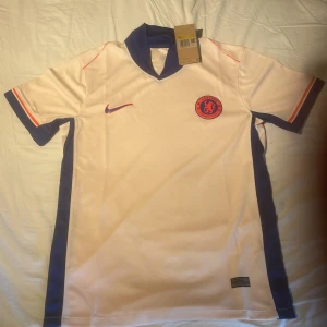 Chelsea FC bortatröja Nike S - Chelsea FC bortatröja från Nike i storlek S. Tröjan är vit med blå och röda detaljer, har korta ärmar och klassisk krage. Chelsea-märket och Nike-loggan syns tydligt på bröstet. Tillverkad i lätt och ventilerande material, perfekt för match eller träning eller Drip!!!!