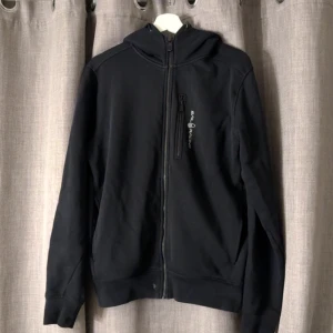 Svart hoodiejacka från Sail Racing M - Om du är intresserad skriv inann du köper! Tyvärr en liten fläck längst ner som ej går bort men annars bra skick! Svart hoodiejacka från Sail Racing med dragkedja framtill och bröstficka med logga. Jackan har huva, ribbade muddar och är tillverkad i mjukt bomullsmaterial. 