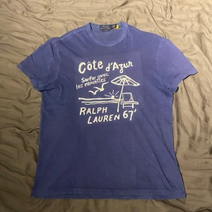  t-shirt Polo Ralph Lauren côte d’Azur blå - Blå T-shirt från Ralph lauren med ett snyggt, sytt motiv på framsida. En tröja som inte längre tillverkas eller säljs med riktigt snygga detaljer så som subtil ”paint splatter” samt aningen urtvättad färg. Bara använts några få gånger och tvättad runt 3-4 gånger. Säljer då den är aningen stor för mig. Skriv om ni undrar nått.                                                          //Hugo