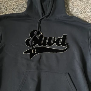 Grå hoodie Pull&Bear med brodyr - Snygg grå hoodie från Pull&Bear med stor svart broderad 'Stwd' logga på bröstet och snörning i huvan. Klassisk känguruficka framtill och mjukt material som känns skönt mot huden. Perfekt för chill dagar eller när du vill ha en avslappnad look. Använd kanske max 3 gånger
