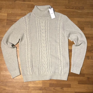 Vit polotröja från Jack & Jones M - Säljer en vitmelerad polotröja från Jack & Jones i storlek M. Tröjan är stickad med flätmönster framtill och har ribbade muddar samt hög polokrage. Perfekt för kalla dagar och riktigt snygg till jeans eller chinos. Helt ny