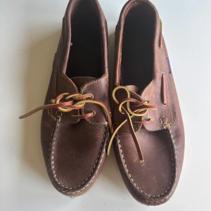 Bruna seglarskor i skinn från Shaka - Klassiska bruna seglarskor från Shaka i äkta skinn med kontrastsömmar och snörning i läder. Skorna har en platt sula i gummi och detaljer i mässing. Perfekta för en avslappnad och stilren look.