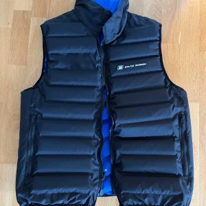 Blå och svart flyväst från Baltic - Säljer en dunväst från Baltic Sweden i blått och svart med quiltad design. Västen har dragkedja framtill, ståkrage och praktiska fickor med dragkedja. Märkeslogga på bröstet och ryggen. Perfekt för dig som vill ha en sportig och snygg look. Går av vända ut och in för att byta färg. Nypris var 1250kr. Helt ny! Aldrig använd med lappen fortfarande kvar. Vikt: 70-80kg