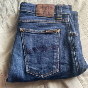 Blå jeans från Nudie Jeans - Snygga blå jeans från Nudie Jeans med klassiska orange sömmar och läderpatch bak i midjan. Jeansen har en rak passform och är tillverkade i slitstarkt denimtyg. Perfekta för dig som gillar en tidlös och stilren look. Har  3 små hål där bak som du kan se på sista bilden 