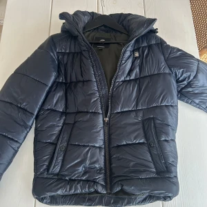 Mörk marinblå pufferjacka från G-Star Raw - Säljer en pufferjacka från G-Star Raw i storlek M. Jackan har huva, dragkedja framtill och två fickor med knappstängning. jackan är perfekt för kalla dagar. Skriv om ni har några frågor. Priset ej hugget i sten