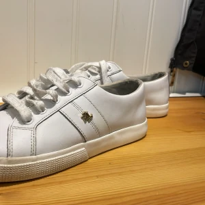 Vita sneakers från Ralph Lauren - Klassiska vita sneakers från Ralph Lauren med guldfärgad logga på sidan. Skorna har snörning, rund tå och är tillverkade i skinn med vit sula. Perfekta för en clean och stilren look.