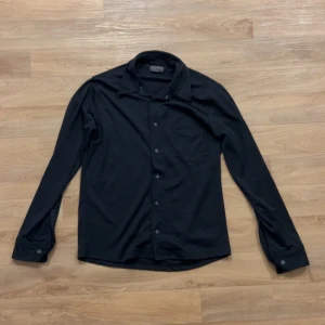 Svart skjorta från Conwell, storlek L - Klassisk svart skjorta från Conwell i storlek L. Skjortan har lång ärm, knappar framtill och en bröstficka. Enkel och stilren design som passar till många olika looks. 