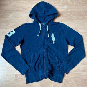 Marinblå hoodie från Polo Ralph Lauren - Säljer en marinblå hoodie från Polo Ralph Lauren med dragkedja, huva och vita snören. Stor broderad logga på bröstet och siffra på ärmen. Mjuk bomullskvalitet och kängurufickor framtill. Perfekt för en chill och sportig look.