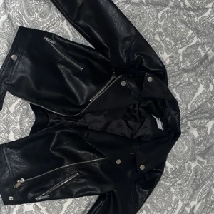 Svart skinnjacka med dragkedjor - Snygg svart skinnjacka med coola silverfärgade dragkedjor och tryckknappar. Klassisk bikerstil med snedställd dragkedja framtill och flera fickor. Perfekt för dig som vill ha en edgy look och gillar tidlös streetstyle.