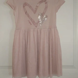 Rosa kortklänning med paljett-hjärtan Lindex 98/104 - Supersöt ljusrosa kortklänning från Lindex med glittrigt hjärta och paljetthjärta framtill. Strl 98/104. Klänningen har korta ärmar och ett mjukt, svängigt kjolparti. Perfekt för dig som gillar glitter och gulliga detaljer. Överlag fint skick, finns ett mini hål vid hjärtat. 