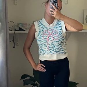 Stickad väst med blått mönster från Zara - Säljer en stickad väst från Zara i vitt med blått zebramönster och en stor rosa blomma på framsidan. Västen är croppad med ribbade kanter vid hals, ärmhål och nederkant. Perfekt att styla över en t-shirt eller skjorta för en cool look.