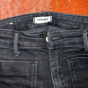 Svarta bootcut jeans från Wrangler - Säljer ett par svarta jeans från Wrangler med bootcut passform och hög midja. Jeansen har framfickor med markerade sömmar och klassiska bakfickor, varav en med Wrangler-logga. Materialet är jeans med stretch och de stängs med knapp och dragkedja.