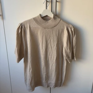 Beige stickad topp från Stockh LM - Snygg beige stickad topp från Stockh LM med korta puffärmar och hög ribbad hals. Materialet är mjukt och har en lätt glansig finish som ger en exklusiv känsla. 