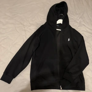 Svart hoodie från polo - Svart hoodie från polo  med klassisk vit logga på bröstet. Tröjan har huva, dragkedja framtill och två fickor. Tillverkad i mjuk bomull, perfekt för chill dagar. Långärmad och med ribbade muddar för extra komfort.