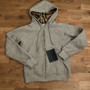 Grå Burberry hoodie med dragkedja - Säljer en grå hoodie från Burberry med klassiskt rutigt foder i huvan och snyggt broderad logga på bröstet. Tröjan har snörning i huvan, dragkedja framtill och ribbade muddar. Perfekt för dig som vill ha en stilren och lyxig look.