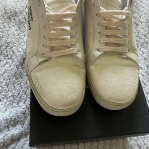 Saint Laurent vita sneakers i canvas - Säljer ett par stilrena vita sneakers från Saint Laurent med handskriven logga på sidan. Skorna har rund tå, platt sula och snörning. Tillverkade i canvas med detaljer i skinn och har en clean, minimalistisk vibe som passar till jeans eller shorts.