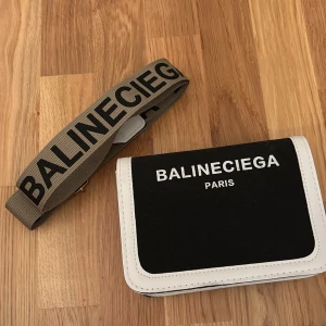 Svart och vit axelväska Balineciega - Snygg axelväska från Balineciega med svart framsida och vita kanter. Väskan har en bred beige axelrem med tydlig logga i svart. Perfekt storlek för mobilen och det viktigaste, med stilren design och tydlig Paris-text framtill.