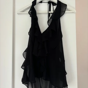 Svart volang topp med knyt i nacken - Säljer en svart, transparent linnetopp med volanger och djup v-ringning. Toppen har knyt i nacken och är ärmlös, vilket ger en snygg och luftig look. Perfekt för dig som gillar att sticka ut med coola detaljer och flowy material.