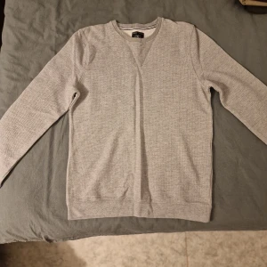 Grå långärmad tröja Hampton Republic - Snygg grå långärmad tröja från Hampton Republic i storlek M. Tröjan har en klassisk rund halsringning och diskret struktur i tyget. Perfekt för dig som gillar stilrena och enkla plagg med en chill vibe. Passar till jeans eller chinos för en clean look.